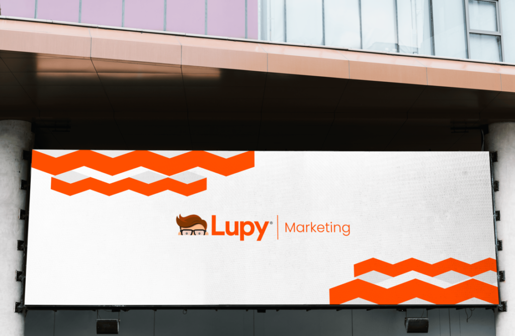 Fachada de um prédio moderno com um grande painel de LED exibindo o logo da Lupy e a palavra "Marketing". O painel tem um fundo branco com detalhes em laranja, incluindo um padrão geométrico na parte superior e inferior. O logo da Lupy apresenta uma ilustração de um personagem com óculos. A imagem destaca a visibilidade e a presença da marca Lupy, sugerindo um espaço comercial ou corporativo dedicado às atividades de marketing da empresa.